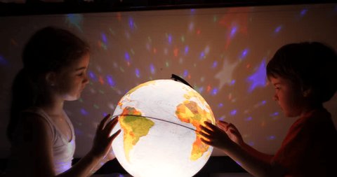 Kids Playing With Globe at Night. Children Sits on Bed in Evening Light Dreaming Vacation. Children Looking at Illuminated Globe, Exploring World, Learning. Dreaming About Future Save of Our Planet.の動画素材