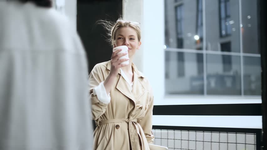 Positive friendly young blond woman hurry to work with cup of hot coffee or tea in the morning and say hi nodding head greeting wave to colleague near modern business centre Dream job concept