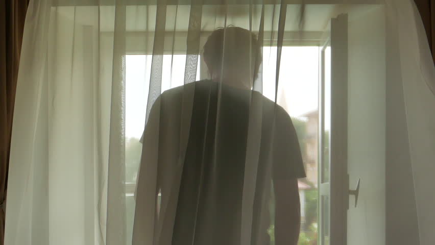 Depressed man at bedroom window behind the curtains looking out and contemplating, rear view, selective focus