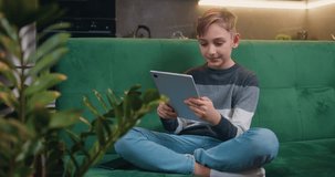 Smiling boy sitting on the couch with his legs crossed holding tablet device, texting message or playing game at home. Leisure, children, technology, internet communication and people concept