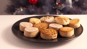 Holiday typical mediterranean delicacies polvorones and mantecados in black plate on white table against backdrop of Christmas Tree.