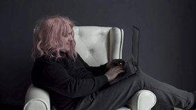 A man in a pink wig sits in a white chair and communicates via video link through a laptop.