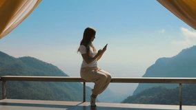Influencer woman sitting on terrace shooting photos or video on mobile phone with beautiful mountain view in background. Life and work of influencers, travel, outdoor lifestyle, camping and vacation