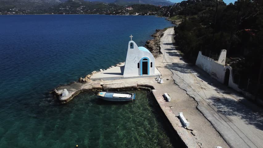 Aerial circular shot over a white little church and a small fishing boat in Lake Vouliagmeni, Loutraki Greece | 4K