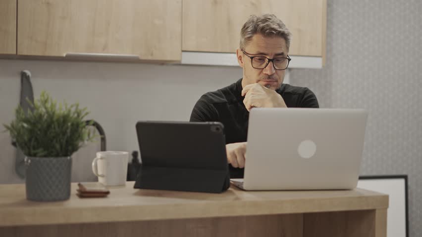 Businessman working with tablet and laptop computer in home office. Happy middle aged, mid adult, mature age man smiling. Entrepreneur sitting at desk in kitchen, managing business online. Royalty-Free Stock Footage #1101915681