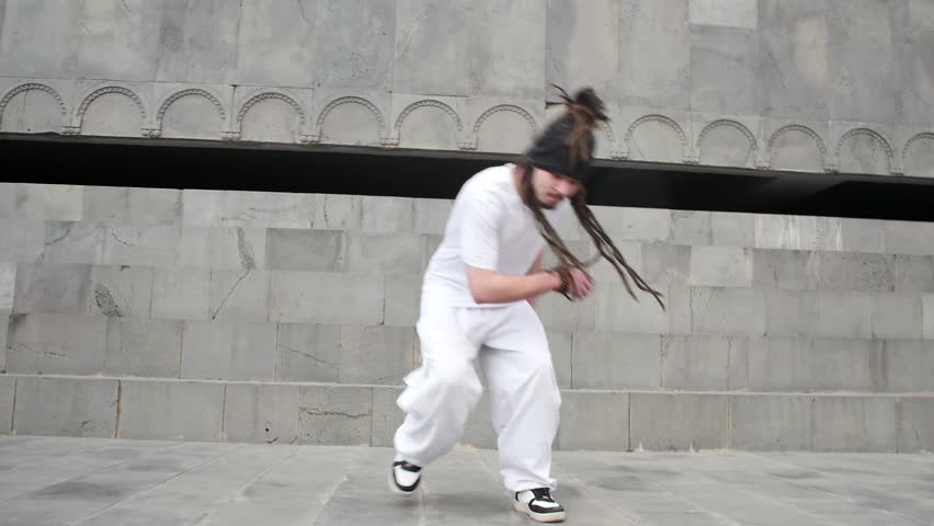 A young handsome man with long dreadlocks on his head in white clothes is dancing capoeira outdoors. Attractive guy practices capoeira brazilian martial art and dance