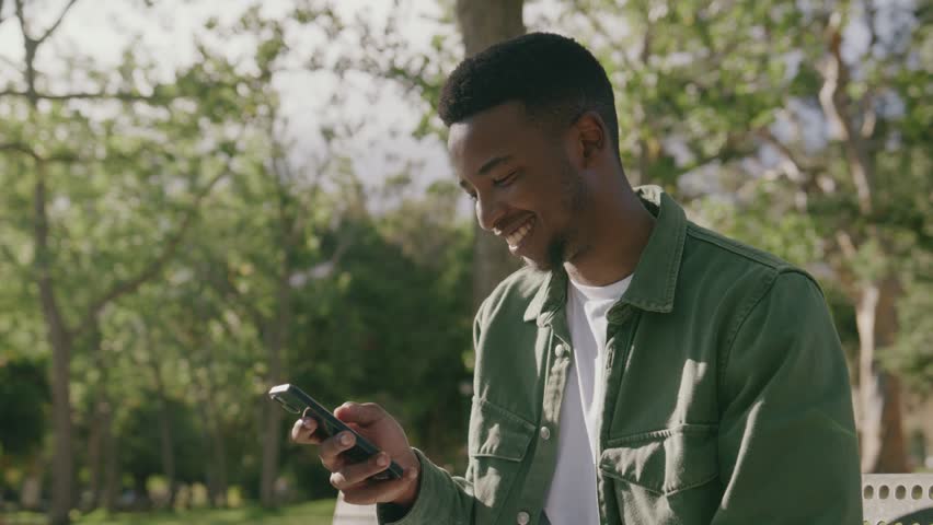 Happy young black man in shirt using mobile phone on bench under tree in park Royalty-Free Stock Footage #1101929105