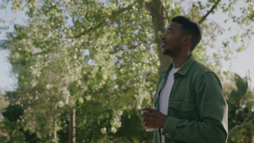 Happy young black man in shirt walking with coffee in disposable cup under trees in park Royalty-Free Stock Footage #1101929113