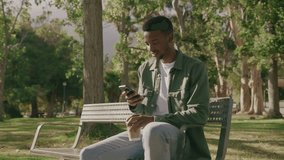 Young black man in shirt using mobile phone and drinking coffee while sitting on bench in park