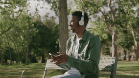 Young black man in casual clothing listening to music on headphones on bench under tree