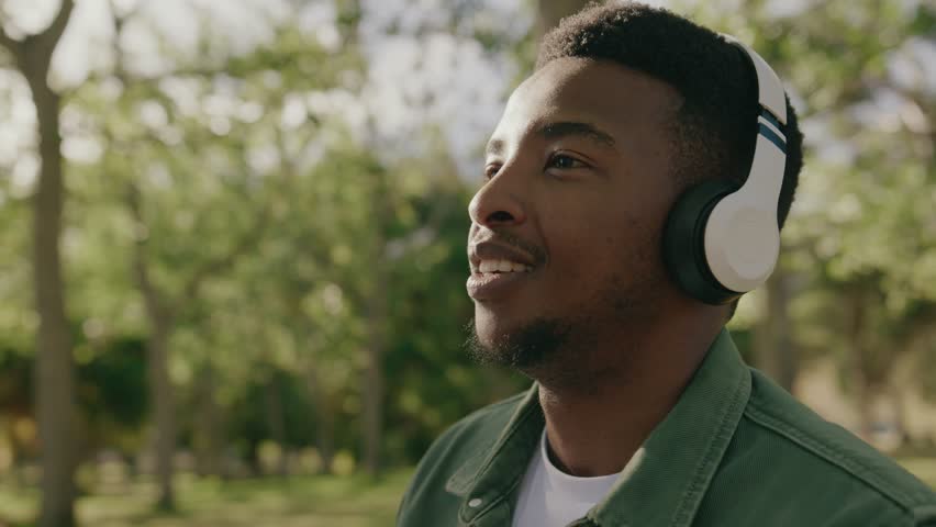 Young black man in shirt smiling and listening to music on headphones under tree in park Royalty-Free Stock Footage #1101929129