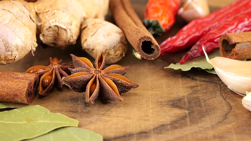 Falling dried clove buds on a wooden board against the background of ginger root, red chili pepper, star anise, bay leaf and garlic.