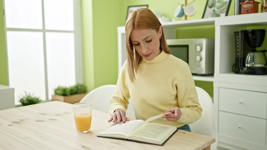 Young blonde woman reading book drinking orange juice at home