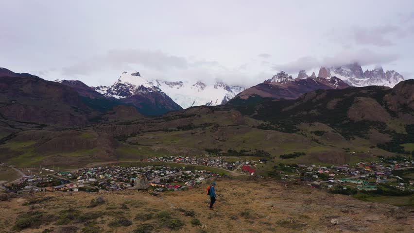Backpacker Traveler Man with Orange Backpack Hiking and Looking at Mount Fitz Roy in Clouds and El Chalten Town. Hills and Mountains. Andes, Patagonia, Argentina. Aerial View. Drone is Panning
