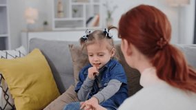 caring mother is interested in health of her cute daughter holding hands while sitting on sofa in cozy room, focus on hands