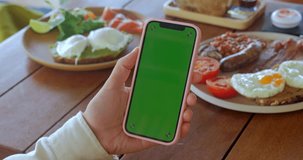 Female hand holding smartphone with green screen chroma key at cafe table with food morning breakfast. Woman holding smartphone in hand with green screen mock up. Order food online with smartphone.