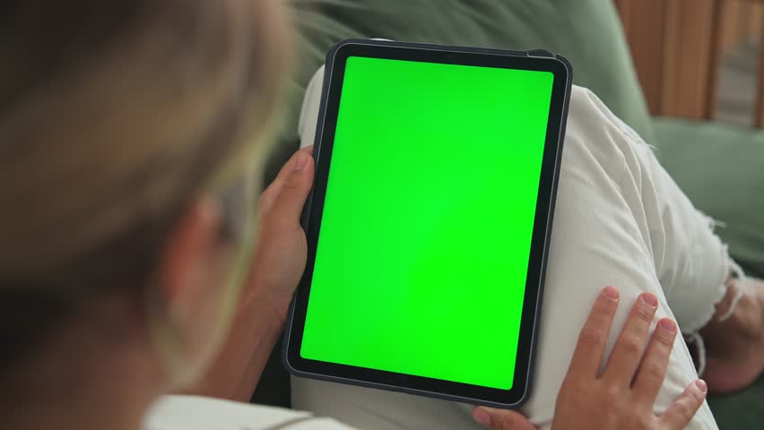 Girl click green screen display. Hand hold vertical ipad close up. Finger tap center tablet job. View green web store site. One touch chroma key pad. Woman poke screens. Person work home room lie sofa