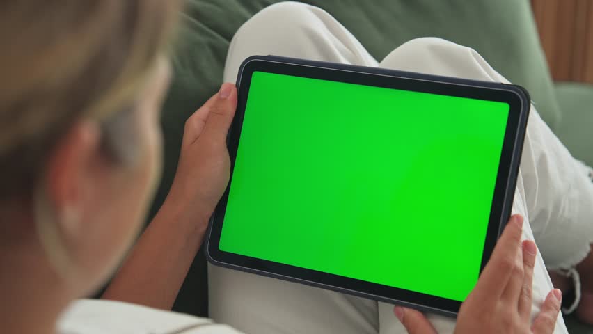 Girl click green screen display. Hand hold horizontal ipad close up. Finger tap center tablet. View green web store site. One touch chroma key pad. Woman poke screens. Person work home room lie sofa.