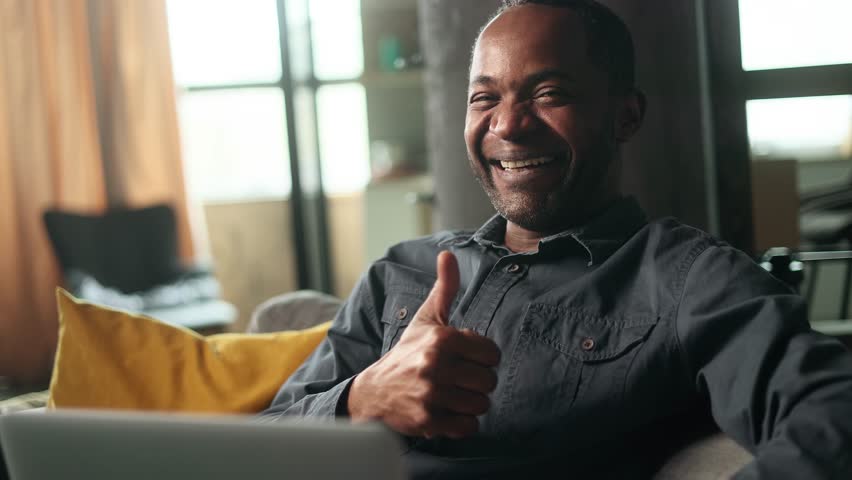 Portrait of cheerful mature african american man looking at laptop screen with nod of approval and looking at camera with thumbs up sitting on sofa at home Advise Approve Recommend Choice concept | Shutterstock HD Video #1101945665
