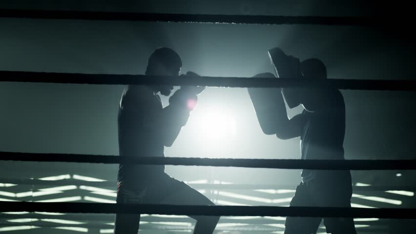 Boxer training and practicing with his trainer in low light gym . Silhouette on dark background. Boxer training in ring . Cinematic slow motion shot of professional young man punching with instructor