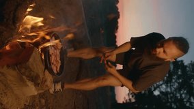 Young male hipster on vacation by the river at sunset roasts meat in pan on fire and turns the pieces with fork. Vertical video