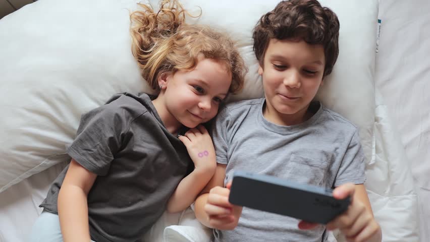 2 children call on the phone on video communication, lying on the bed, top view, close up. The concept of children alone at home