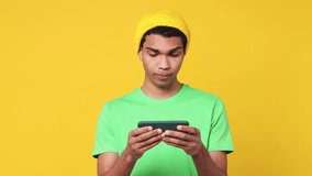Gambling excitable young man of African American ethnicity wear green t-shirt hat use play racing app on mobile cell phone hold gadget smartphone for pc video games isolated on plain yellow background