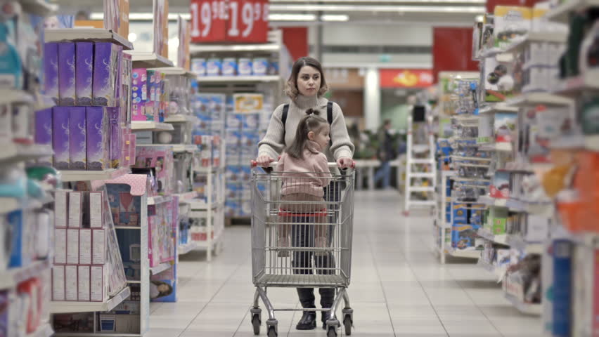 Beautiful young woman is walking through a supermarket with a cart in which a little girl is sitting.