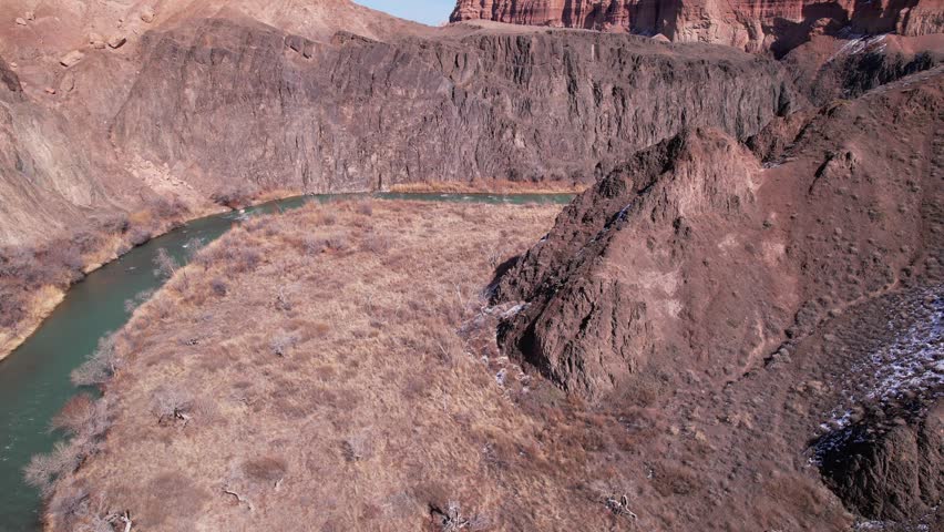 A long river with green water in the Charyn Canyon. There is white snow in places. Bushes and trees grow. The river runs through a canyon among rocks and cliffs. Mountains are visible. Drone view