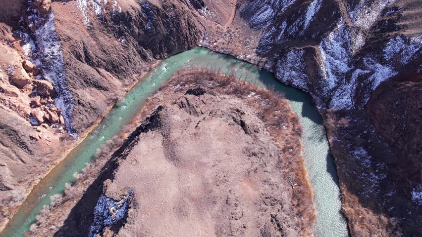 A long river with green water in the Charyn Canyon. There is white snow in places. Bushes and trees grow. The river runs through a canyon among rocks and cliffs. Mountains are visible. Drone view