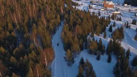 Top view of snowmobile and village in forest in winter. Clip. Cinematic view of snowmobile going to old village. Snowmobile in forest on background of village and mountains in winter