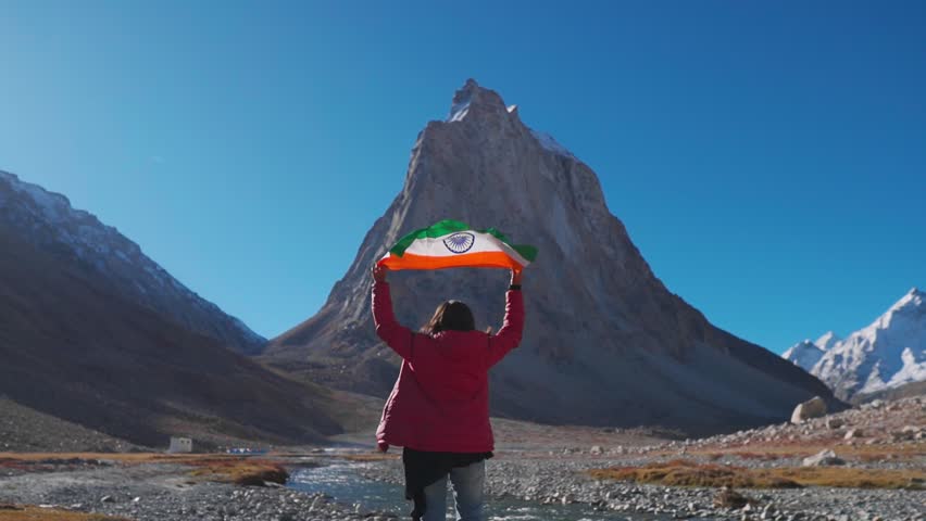 Wide angle shot of female Indian traveler holding Indian flag above her head in front of Gumbok Rangan mountain at Zanskar, Ladakh, India. Indian flag waving in the wind. Indian flag background