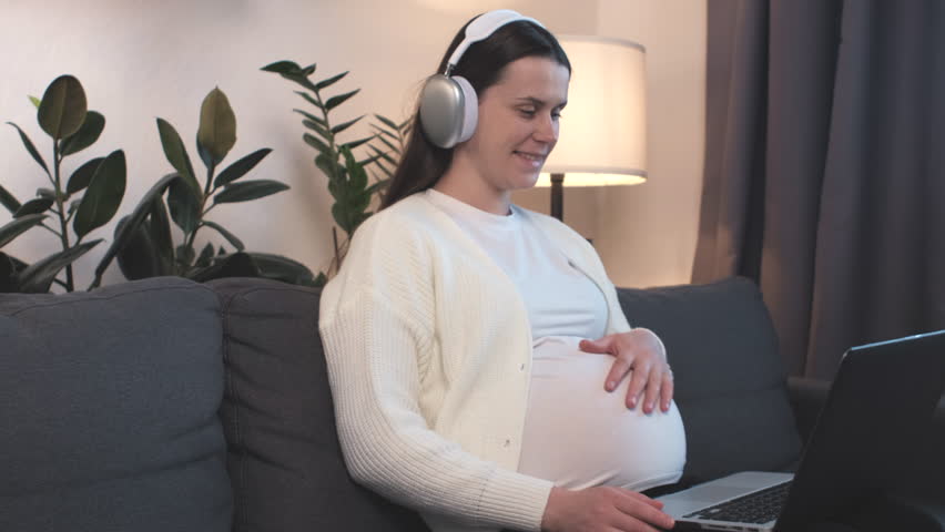 Cute pregnant woman in headphones sit on couch using laptop having video call conversation to family. Future mom talk to obstetrician-gynecologist, receive professional medical online consultation | Shutterstock HD Video #1101974385