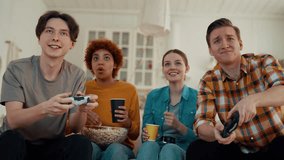 Company of multiethnic friends play video games at home. Two guys competing and two girls cheering them drinking and eating. Diverse people having fun hanging together. Leisure, entertainment concept.