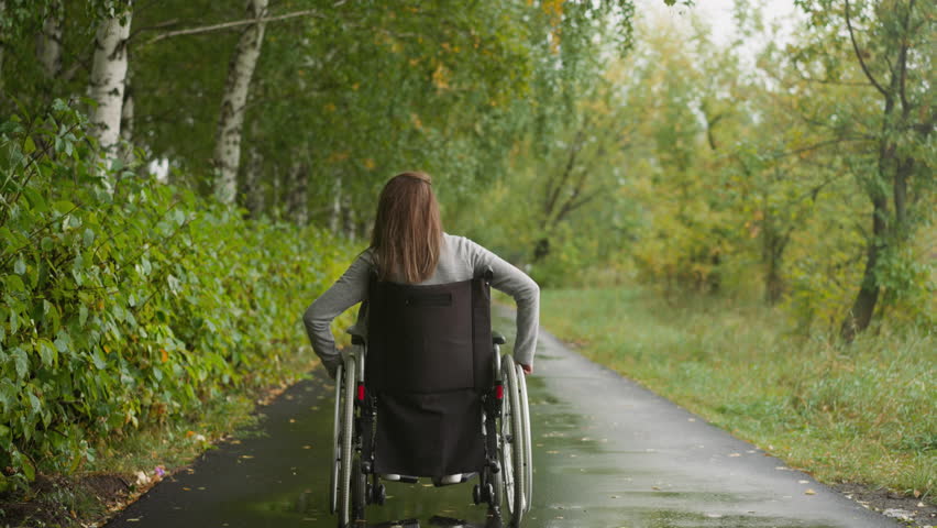 Young woman in wheelchair walks in park in drizzling rain. Female spins wheels moving along wet path. Physical rehabilitation after injury backside view