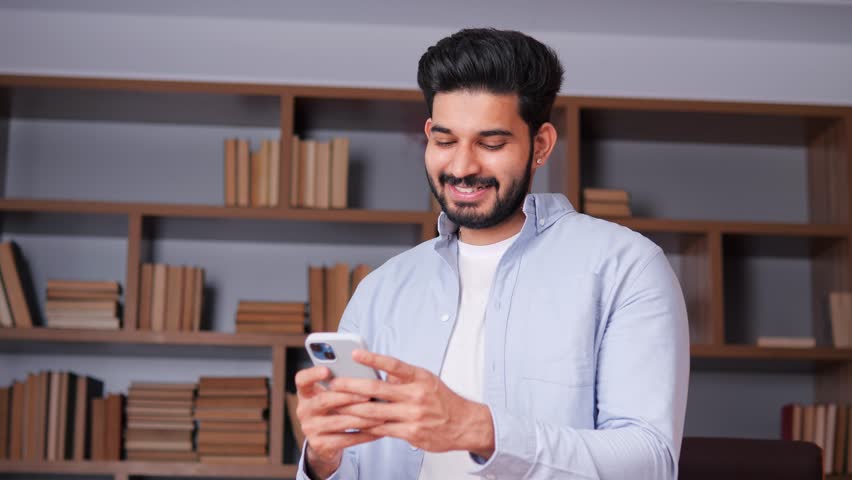 Happy Indian businessman in an office environment, tapping and scrolling on his mobile device while appearing to be deep in thought. Concepts related to mobile technology, communication, productivity.