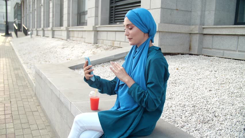 Arab female and technologies concept.. Young muslim woman talking on video call outdoors