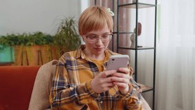 Portrait of young woman sitting on sofa uses mobile phone smiling at home living room apartment. Girl texting share messages content on smartphone social media applications online watching relax movie