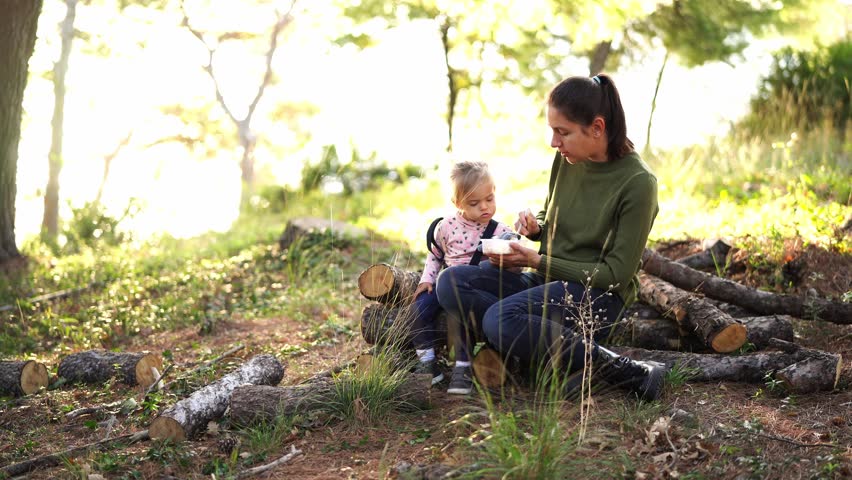Mom feeds a little girl from a spoon sitting on stumps in the park