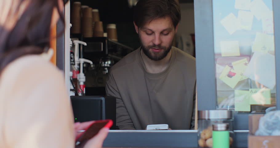 Barista man makes takeaway coffee for a woman customer which pays by contactless mobile phone to credit card system. Royalty-Free Stock Footage #1101984431