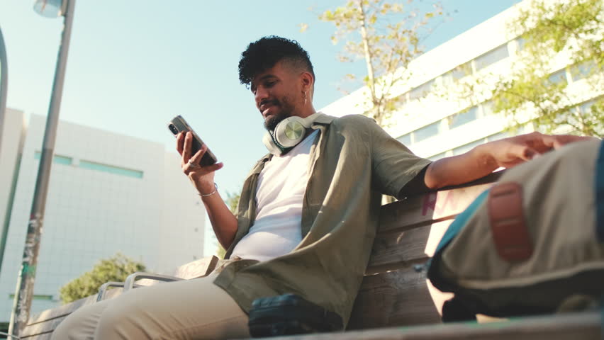 Young man with beard wearing an olive-colored shirt with headphones sits on bench and uses cellphone