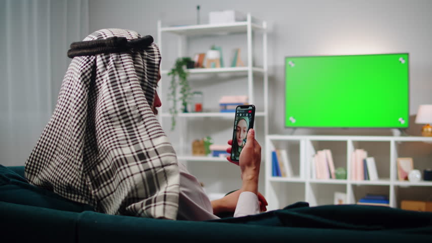 Middle eastern man talking on video call, virtual conference on smartphone. Television with green chroma key screen in living room. Wearing traditional Islamic male clothes. | Shutterstock HD Video #1102002169