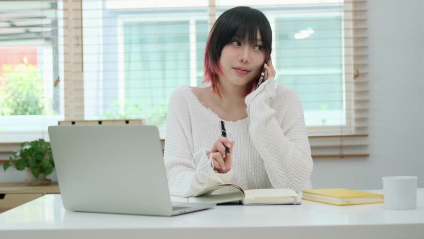 Pretty young creative woman having pleasant phone conversation and making notes on notebook. | Shutterstock HD Video #1102006825