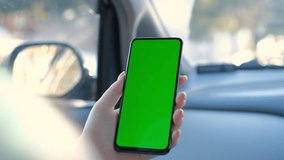 A hand holding a smartphone with green blank screen in the car for direction, massage, location, business. Woman sits in modern car and works on smartphone - green screen - closeup. Chroma key