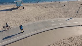 aerial footage of an African American man riding a OneWheel electric skateboard on a bike path with ocean water, brown sand, blue sky at Belmont Shore Beach in Long Beach California