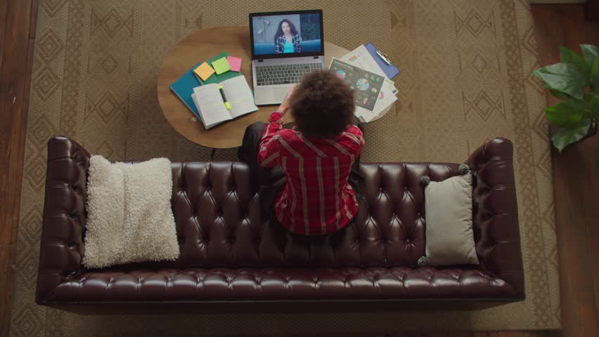 Overhead view of positive charming black young woman with curly hair sitting on couch, friendly chatting online with friend on video call using laptop pc while enjoying leisure indoors. | Shutterstock HD Video #1102010149