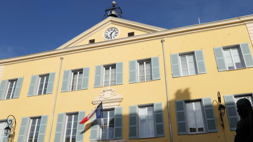 French flag flies on the newly painted facade of Antibes town hall - Hotel de Ville (Mairie).  Sunny day with clear blue Spring sky. French Mediterranean Antibes sits between Nice and Cannes. Royalty-Free Stock Footage #1102014001