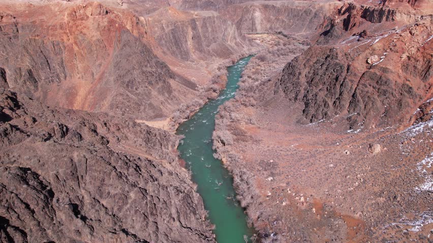 A long river with green water in the Charyn Canyon. There is white snow in places. Bushes and trees grow. The river runs through a canyon among rocks and cliffs. Mountains are visible. Drone view