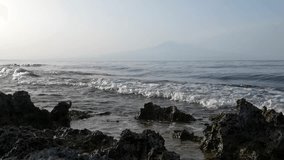 Waves breaking on the rocks at sunset. Etna volcano in the background. Super slow motion