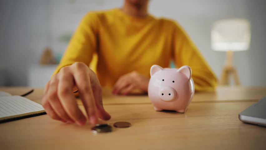 African american woman in yellow sweatshirt puts coins in pink piggy bank sits at desk at home in modern interior, hand close-up. Saving money, self finance and savings capital concept. | Shutterstock HD Video #1102019599
